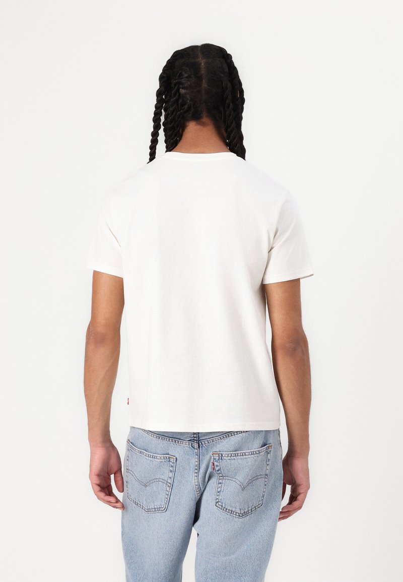 Cremefarbenes Kurzarm-T-Shirt mit Rundhalsausschnitt, figurbetontem Schnitt und sichtbarem rotem Etikett auf der linken Seite. Getragen mit blauen Jeans.