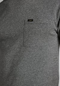 Tummanharmaa, teksturoidusta kankaasta valmistettu t-paita, jossa on pieni suorakaiteen muotoinen tasku vasemmalla rinnalla, jossa musta-keltainen "Lee"-lappu.