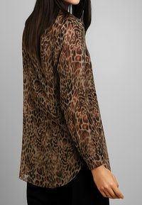 Blusa trasparente marrone con motivo leopardato, maniche lunghe e orlo arrotondato. Il tessuto appare leggero con una texture liscia.