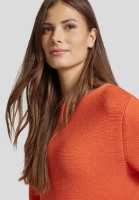 Texturiertes orangefarbenes Strickpullover mit rundem Halsausschnitt, lockerer Passform und langen Ärmeln, getragen von einer Person mit langen, braunen Haaren.