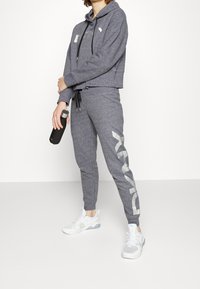 Šedá zkrácená mikina s kapucí a šňůrkou, v kombinaci s odpovídajícími tepláky s velkým potiskem loga. Bílé sportovní boty a černá láhev na vodu.