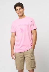 Rožinė medvilninė marškinė su apvaliu kaklu ir trumpomis rankovėmis, su U.S. Polo Assn. logotipu šviesesnėje rožinėje spalvoje. Derinamos su smėlio spalvos krovinių šortais.