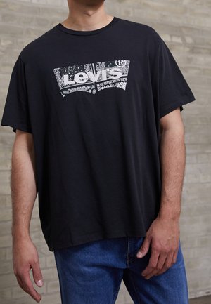 Mężczyzna noszący czarną koszulkę Levi’s z białym logo paisley i niebieskimi dżinsami, stojący na tle jasnej, ceglanej ściany.