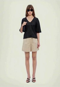 Vrouw in een zwarte glinsterende V-hals top, beige knielange shorts, zwarte sandalen, met een zwarte handtas en donkere zonnebril.