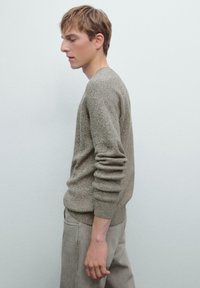 Maglione in maglia marrone chiaro con polsini a costine e scollatura tonda, abbinato a pantaloni grigi. Materiale texturizzato e design aderente.