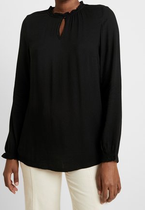 Frau trägt eine schwarze Bluse mit langen Ärmeln, Rüschen am Kragen und an den Manschetten, kombiniert mit hellen Hosen, vor einem schlichten Hintergrund.