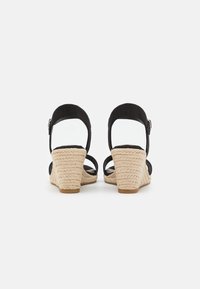 Un paio di sandali neri in suede con zeppa in paglia intrecciata e cinturini alla caviglia, mostrati da dietro su uno sfondo bianco.