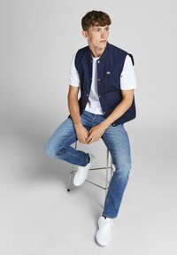 Ναυτικό αμάνικο μπουφάν πάνω από άσπρη μπλούζα, συνδυασμένο με ανοιχτό μπλε τζιν και άσπρα αθλητικά παπούτσια. Διαθέτει κουμπιά και δύο μπροστινές τσέπες.