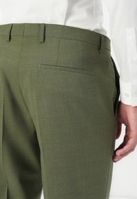 Olivgrüne maßgeschneiderte Hose mit feiner Textur, ausgestattet mit einer Gesäßtasche und Gürtelschlaufen. Der Stoff wirkt glatt und strukturiert.