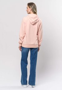 Svetlo roza kapucar z mehko teksturo, z zavihanimi rokavi in žepom kenguru ter modrimi širokimi kavbojkami.