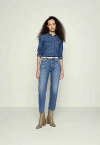 Mulher com cabelo longo e encaracolado, vestindo uma camisa de ganga, jeans de cintura alta, um cinto branco e botas de tornozelo bege, de pé contra um fundo simples.