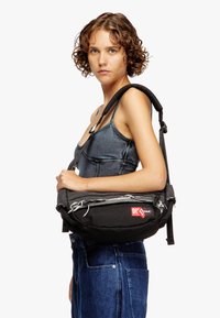Black canvas shoulder bag with frayed edges, adjustable strap, and red logo patch. Compact design with a textured finish.