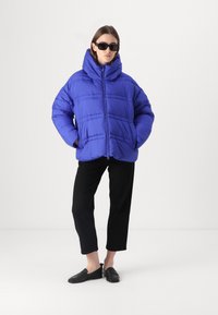 Königsblauer Daunenjacke mit hohem Kragen, Reißverschluss und gestepptem Design. Kombiniert mit schwarzen, knöchellangen Hosen und schwarzen Slippern.
