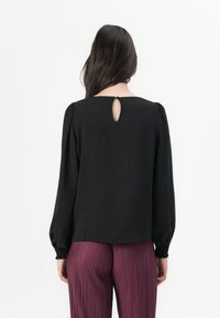 Svart bluse med lange, puffede ermer, detalj med nøkkelhull bak og teksturert stoff; parret med plisserte burgunder bukser.