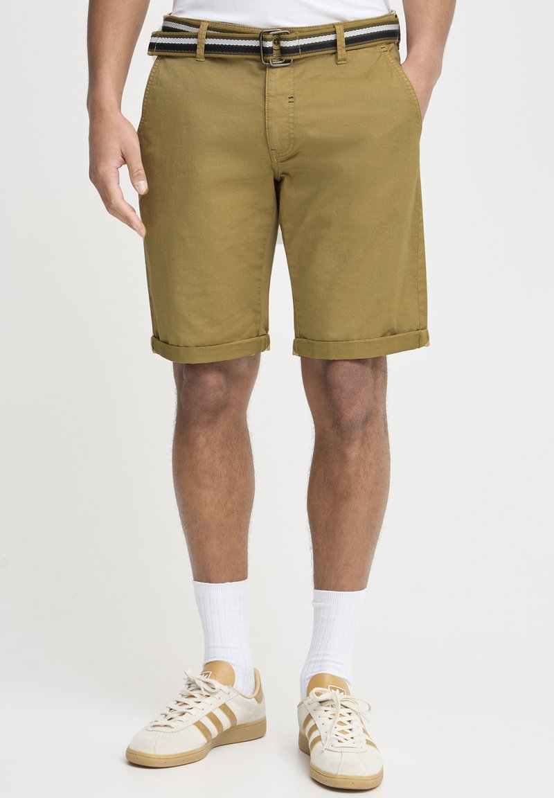 Homme portant un short kaki retroussé avec une ceinture rayée, des chaussettes blanches et des baskets beige et blanches, debout devant un fond uni.