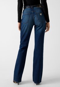 Jeans a zampa in denim blu scuro con vita alta, cinque tasche e un piccolo logo triangolare sulla tasca posteriore destra. Texture liscia.