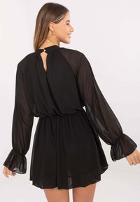 Kobieta uśmiechająca się, ubrana w czarną sukienkę z długim rękawem z przezroczystymi rękawami, elastycznym pasem i małym zapięciem na guzik z tyłu przy dekolcie.