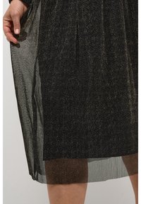 Gonna plissettata nera e oro con sovrapposizione trasparente, caratterizzata da una texture luminosa e un design fluido. Orlo leggermente traslucido.