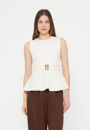 Femme portant un haut peplum blanc sans manches avec boucle de ceinture, associé à un pantalon marron ample, sur fond blanc uni.