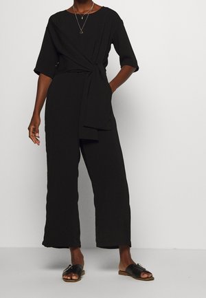 Combinaison noire avec une ceinture à nouer à la taille, manches courtes et jambes larges. Portée avec des sandales plates noires, présentant un accent métallique sur la bride.