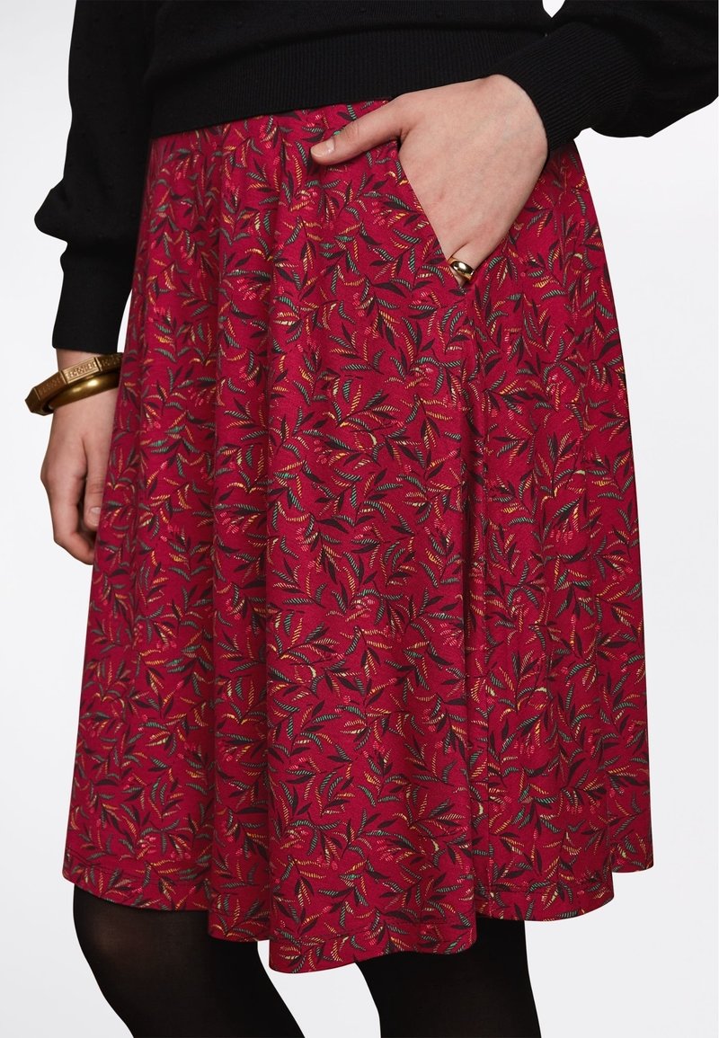 Jupe rouge avec un motif de feuilles dans différentes couleurs. Fabriquée à partir d'un tissu texturé, elle dispose d'une poche latérale et d'une coupe évasée.