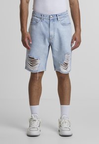 Lyseblå denimshorts med slidte huller, frynset kant, fem lommer og en afslappet pasform. Båret sammen med hvide sneakers og sokker.