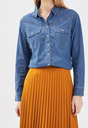 Femme portant une chemise en denim bleu à manches longues avec des poches poitrine boutonnées et une jupe plissée jaune moutarde taille haute.