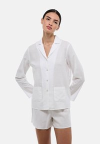 Chemise en lin blanche à manches longues avec col, deux poches avant, associée à un short blanc assorti.