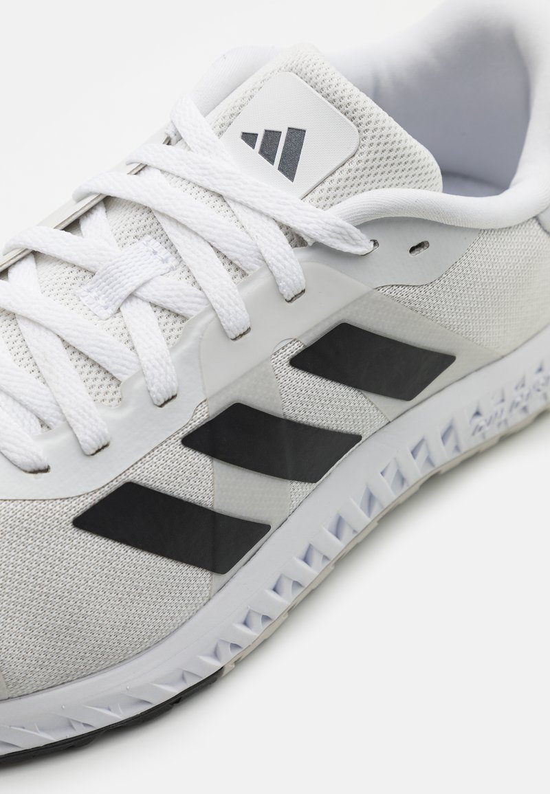 Weißer Sportschuh mit einem Mesh-Obermaterial, drei schwarzen Streifen an der Seite und einer strukturierten Gummisohle. Runde Schnürsenkel und gepolsterter Schaft.