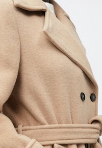 Beige gyapjú kabát dupla gombolású kialakítással, kivágott gallérral, fekete gombokkal és szövet övvel. Puha tapintású, meleg megjelenésű.
