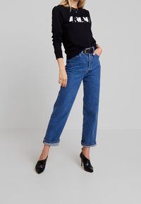 Svart sweatshirt med vit logotyp, blå högmidjade jeans med uppvikta fållar och svarta spetsiga klackskor. Slät textur överallt.