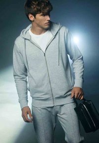 Szary zamek z kapturem z przednią kieszenią, zestawiony z dopasowanymi dresami. Model trzyma małą czarną torbę, prezentując swobodny strój sportowy.