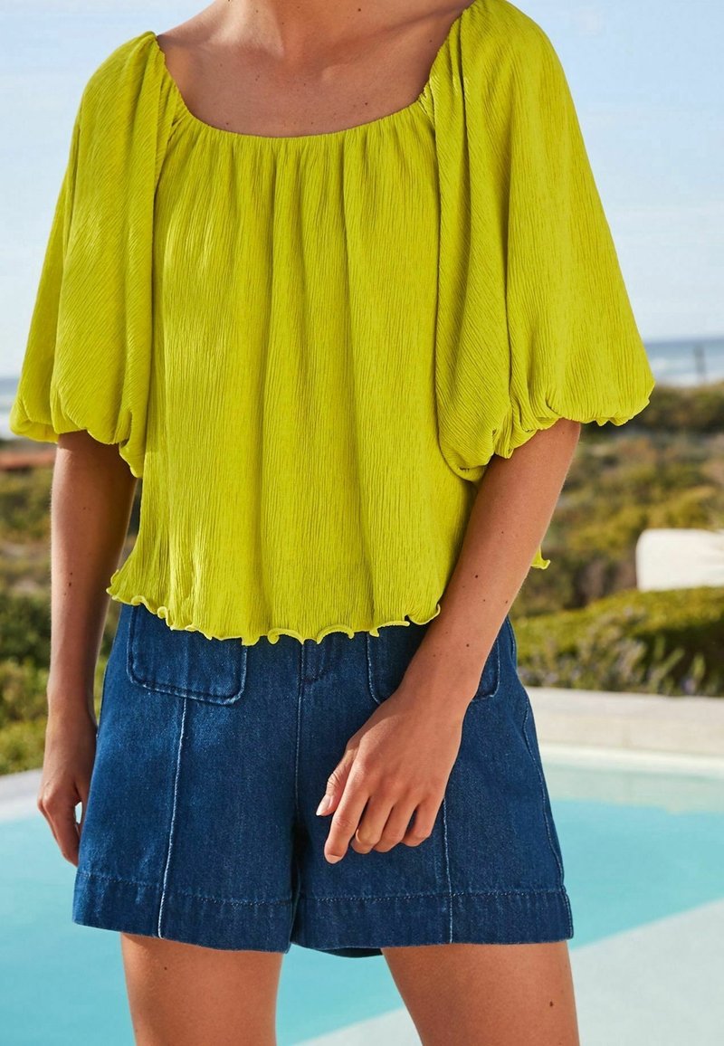 Blouse vert citron texturée avec manches bouffantes, ourlet festonné, associée à un short en jean foncé avec deux poches avant.