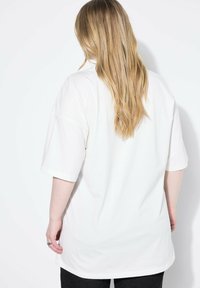Hvid oversized T-shirt med korte ærmer, glat tekstur, rund kant og en afslappet pasform, båret med mørke bukser, vist fra ryggen.