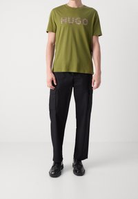Olive green t-shirt z krótkim rękawem z napisem "HUGO" w czarnym kolorze, w połączeniu z czarnymi spodniami cargo z bocznymi kieszeniami i czarnymi butami.