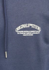 Sudadera color navy con un tejido texturizado, que presenta el texto bordado en blanco "ORIGINALS™ STUDIO" y un cordón plateado con tope.