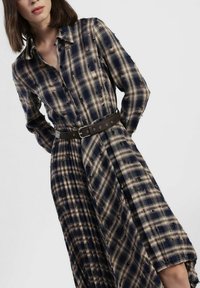 Lungo vestito a quadri in blu navy e beige, con chiusura frontale a bottoni e vita cinturata. Realizzato in tessuto morbido con vestibilità comoda.