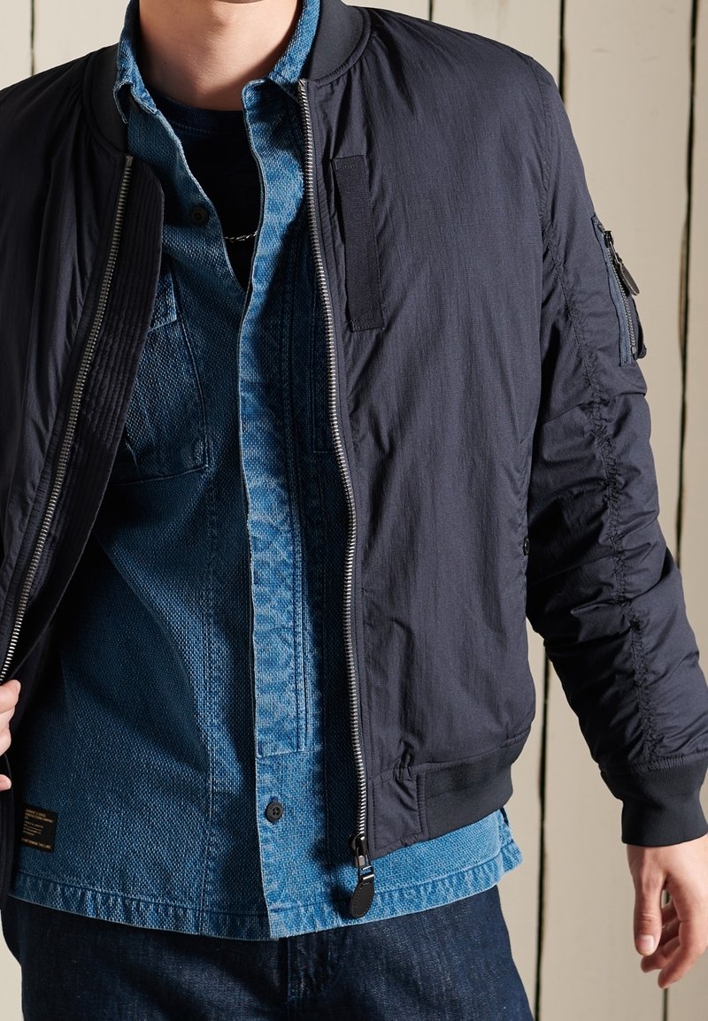Navy bomber jacket with ribbed cuffs and collar, featuring zip closure and textured sleeves. Worn over light blue denim shirt.