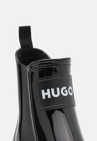 Botín negro brillante hasta el tobillo con panel lateral elástico y lengüeta, marcado con el logo "HUGO" en la parte delantera.