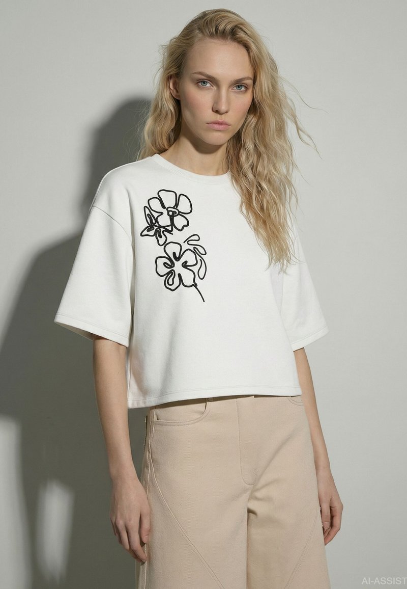 Femme blonde aux cheveux ondulés portant un haut blanc à manches courtes avec un motif floral noir et un pantalon beige taille haute sur fond gris.