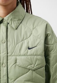 Jachetă puf de culoare verde, cu un guler ascuțit, nasturi cu prindere tip snap și un logo black Nike pe piept. Material moale și texturat.