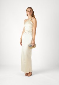 Satin haljina za večer u off-white boji, s jednim naramenicom i prilagođenim siluetom. Uparena s metalnom clutch torbicom i sandalama sa remenčićima.