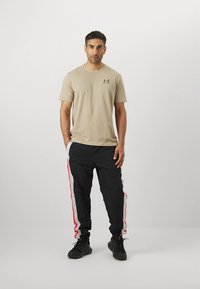 Camiseta bege de algodão com mangas curtas e logótipo no peito, combinada com calças pretas com riscas vermelhas laterais e ténis pretos.