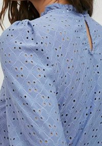 Vaaleansininen paita, jossa on tekstuuripintainen kangas, brodeerattuja kuvioita ja aukkoja. Röyhelöpääntie ja avattava selkäosa.