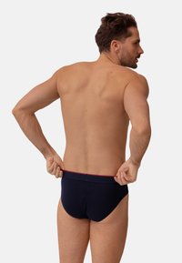Slip blu navy con cintura rossa, caratterizzato da una texture liscia e un design a vita bassa, mostrato da dietro con un modello che lo aggiusta.