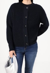 Marinblå knapp-up kardiagan tillverkad av strukturerad stickad material med långa ärmar och framknappar; i kombination med blå jeans och en silverhandväska.