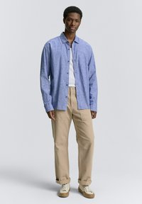 Lichtblauw, knoopsluiting overhemd van geweven stof, gedragen over een wit T-shirt. Gecombineerd met beige, losse broek en beige sneakers.