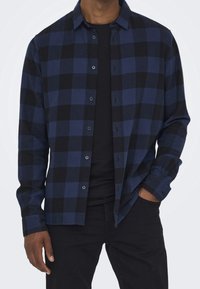 Mężczyzna ubrany w niebiesko-czarną kraciastą koszulę na guziki, na czarnym T-shircie, w czarnych spodniach, z jedną ręką w kieszeni.