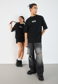 Black oversized t-shirts with white graphic print, paired with distressed gray denim and black sneakers. Model poses against a plain wall.