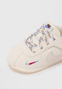 Krupan prikaz bež Nike tenisice s dušicama u više boja i malim izvezenim Nike ševronom u crvenoj, plavoj i svijetloplavoj boji blizu prsta.